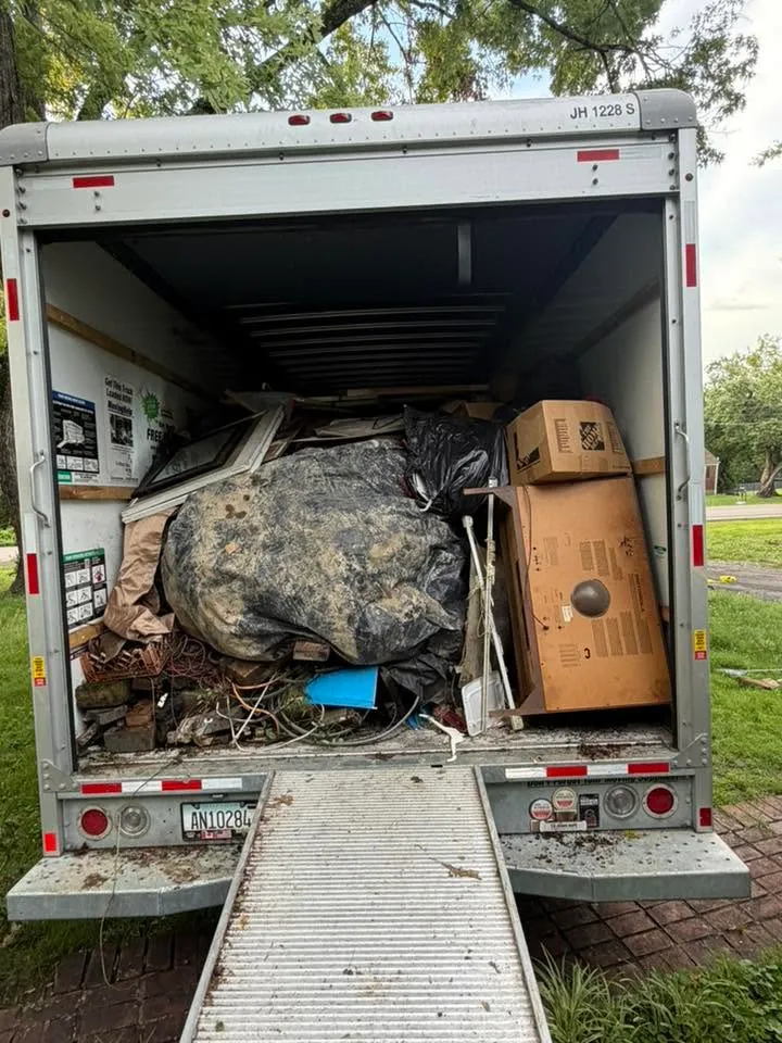 A box truck backed up with its ramp down, the bed packed with a bundled tarp, a dresser and household junk