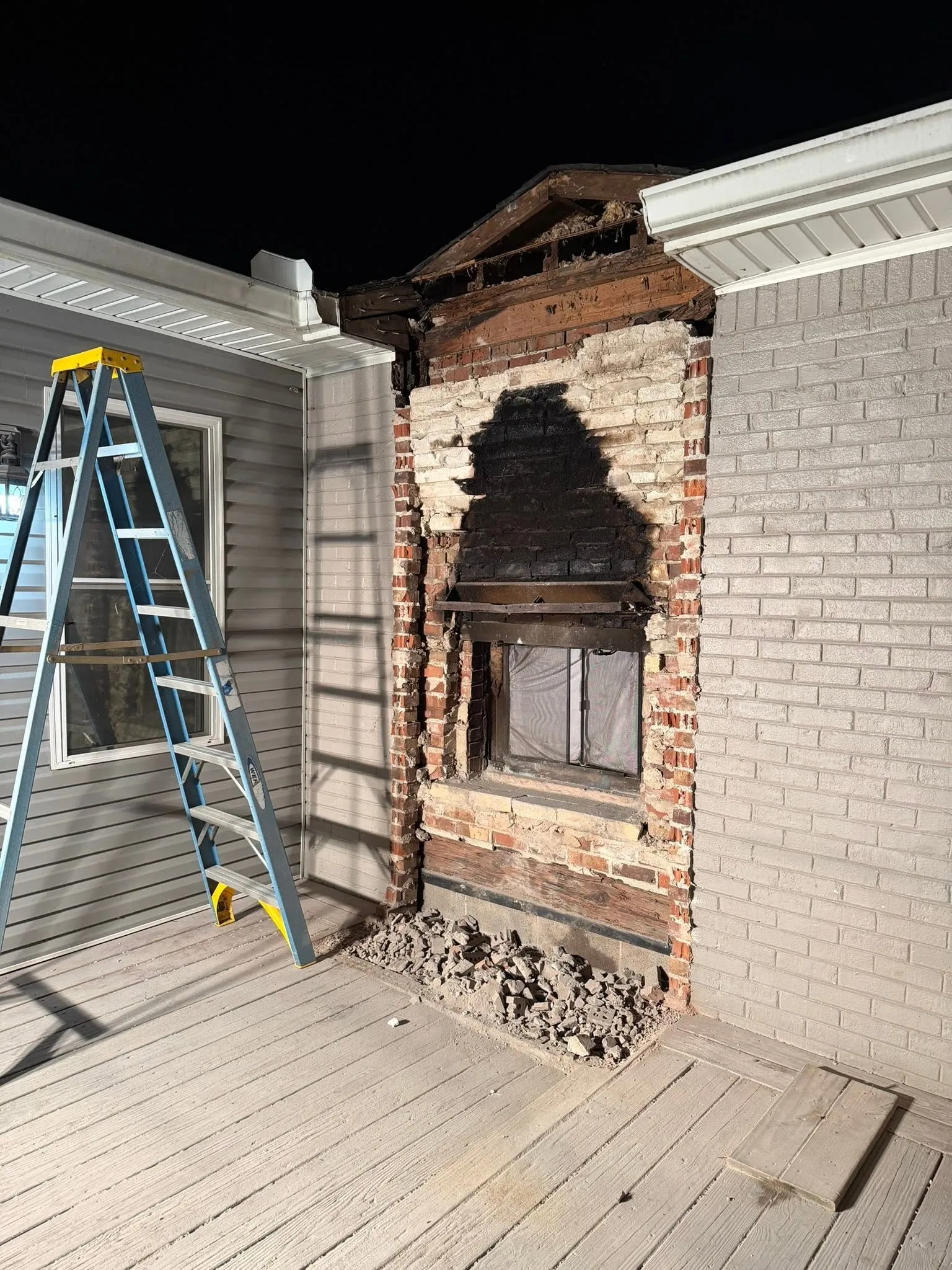 A brick fireplace partly torn down on a covered deck at night, a ladder beside it and rubble at its base