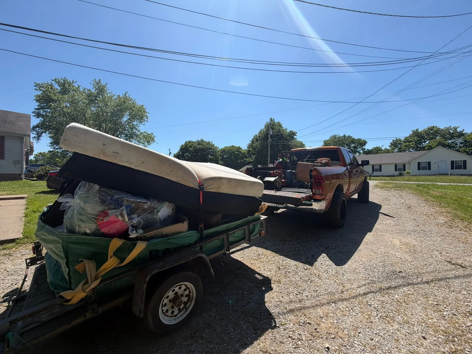 The orange pickup towing a trailer stacked with a mattress, boxes and junk down a residential street