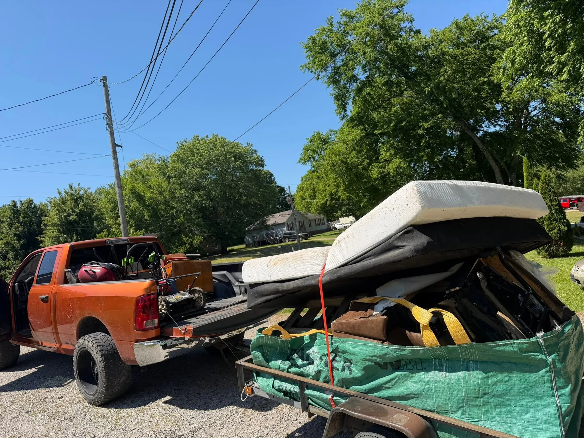 The orange pickup and its trailer stacked high with mattresses, a box spring and bagged junk, parked on a gravel lot