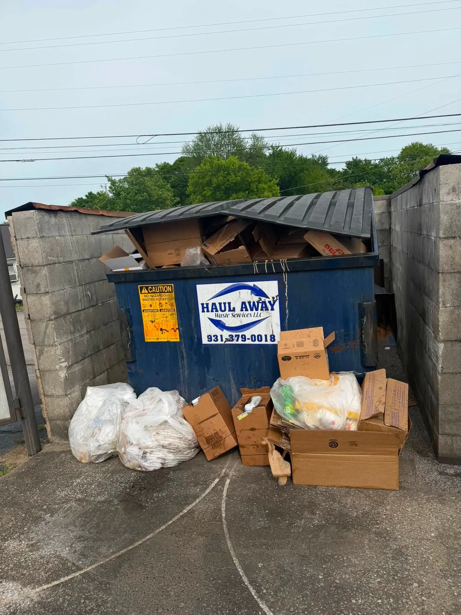 A blue commercial dumpster behind a store, overflowing with boxes and bags stacked beside it