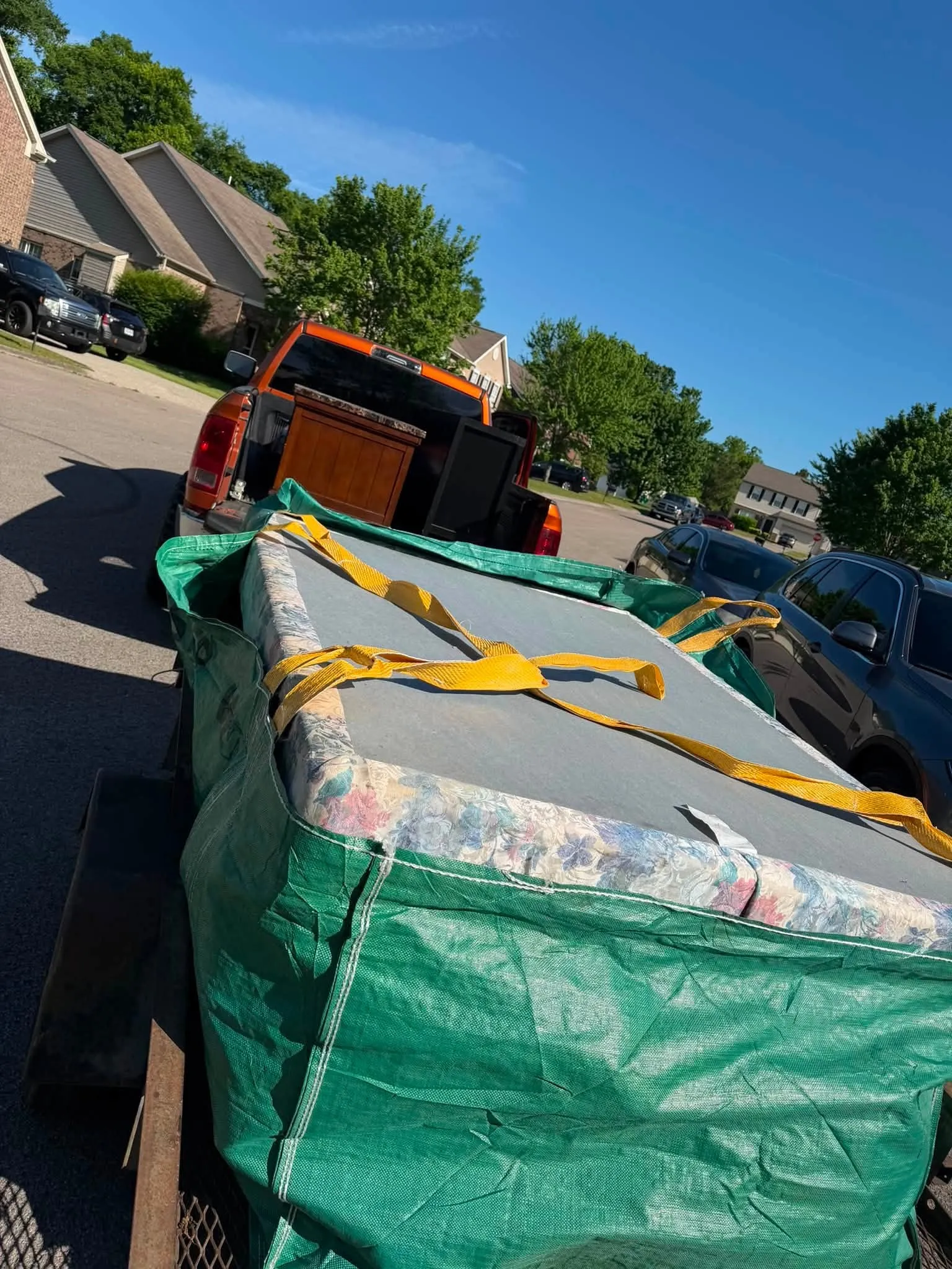 A trailer load strapped down under a green tarp, hitched to the orange pickup in an apartment lot