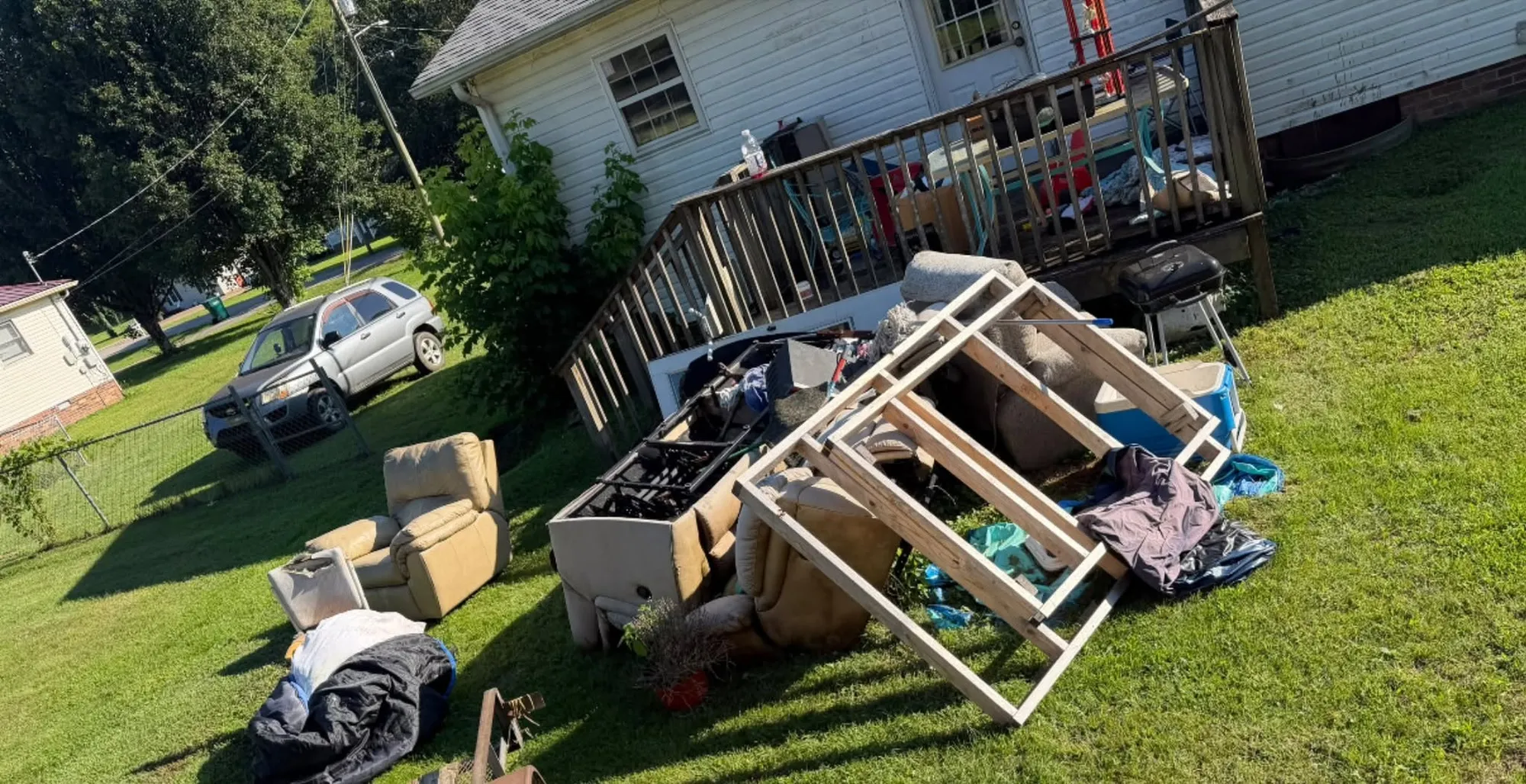A side yard with a wooden shelf unit, boxes and household junk piled on the grass beside a deck